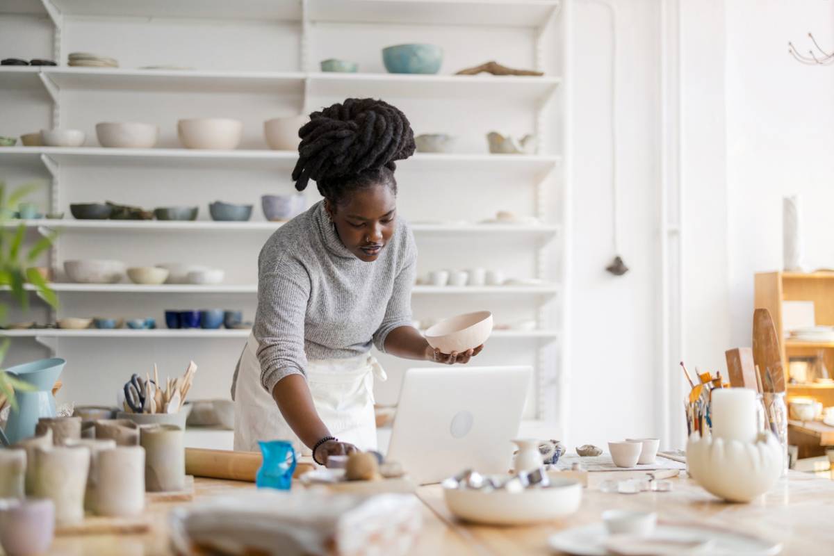 Femme travaillant dans un studio de céramique avec laptop, entourée de poteries blanches, d'outils et d'étagères remplies de bols et tasses.