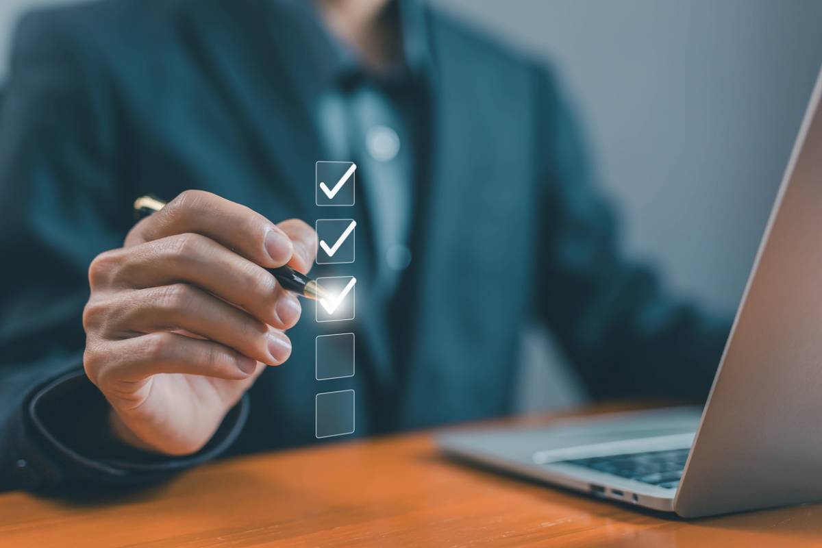 Personne en chemise bleue pointant vers une liste de contrôle numérique avec cases à cocher, devant un ordinateur portable sur un bureau en bois.