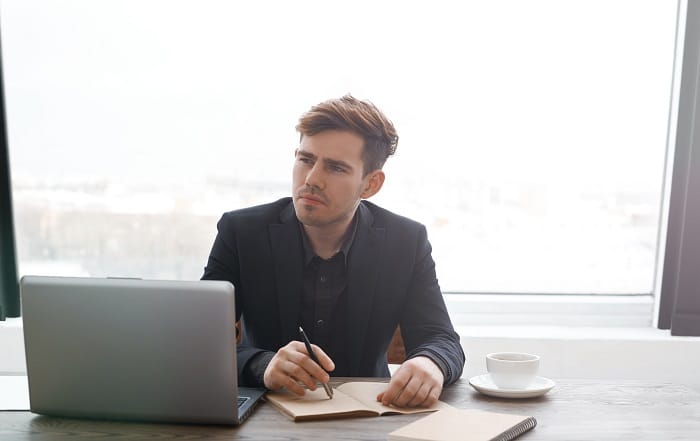 Homme en costume noir travaillant à son bureau avec un ordinateur portable, prenant des notes pour son bilan de compétences, près d'une grande fenêtre.
