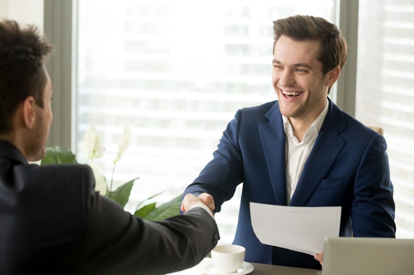 Deux professionnels en costume se serrent la main dans un bureau moderne avec vue sur la ville, l'un tenant un document.