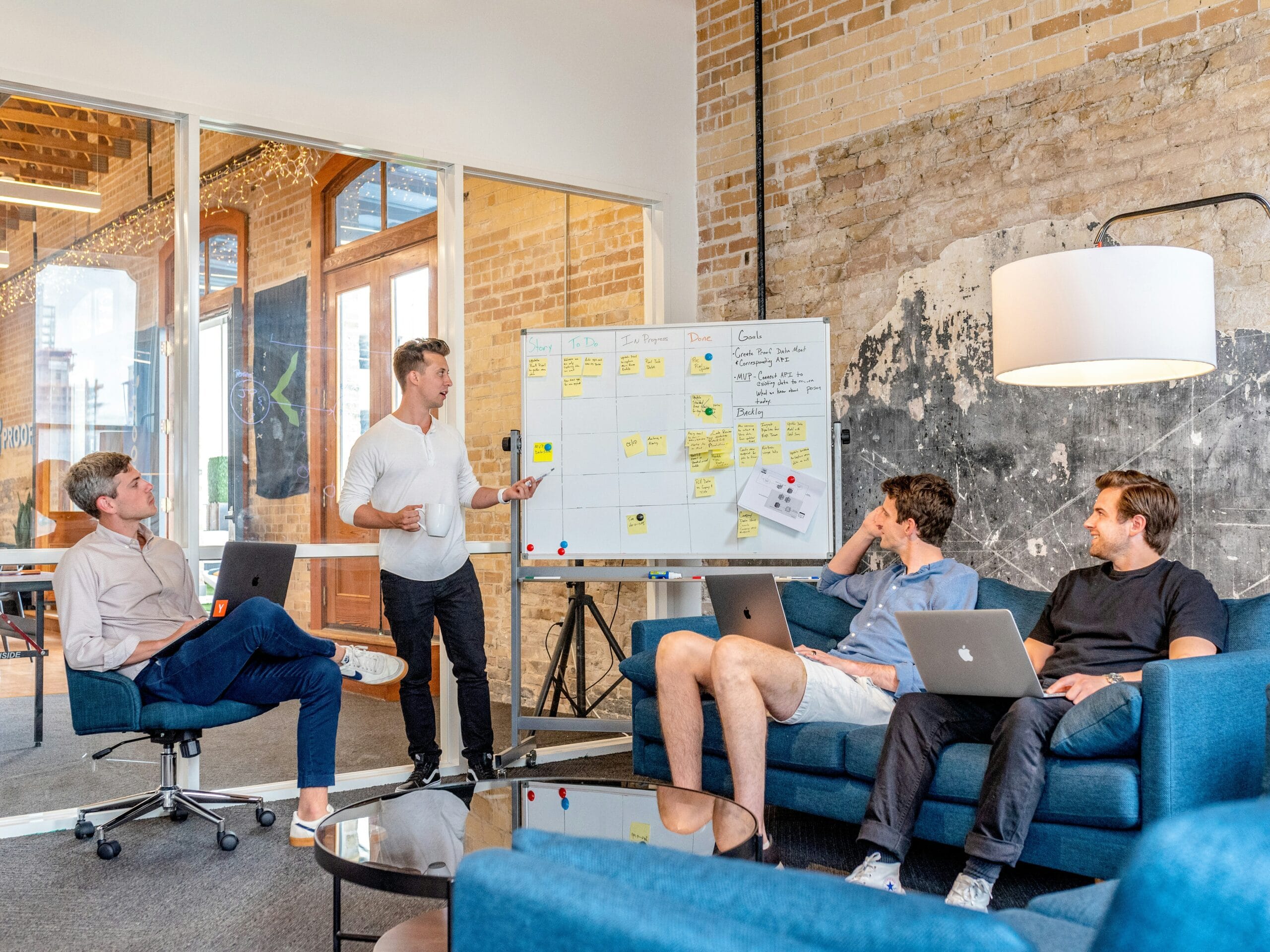 Équipe de quatre hommes discutant autour d'un tableau blanc avec post-its, dans un espace de travail moderne avec briques apparentes, canapé bleu et ordinateurs portables.