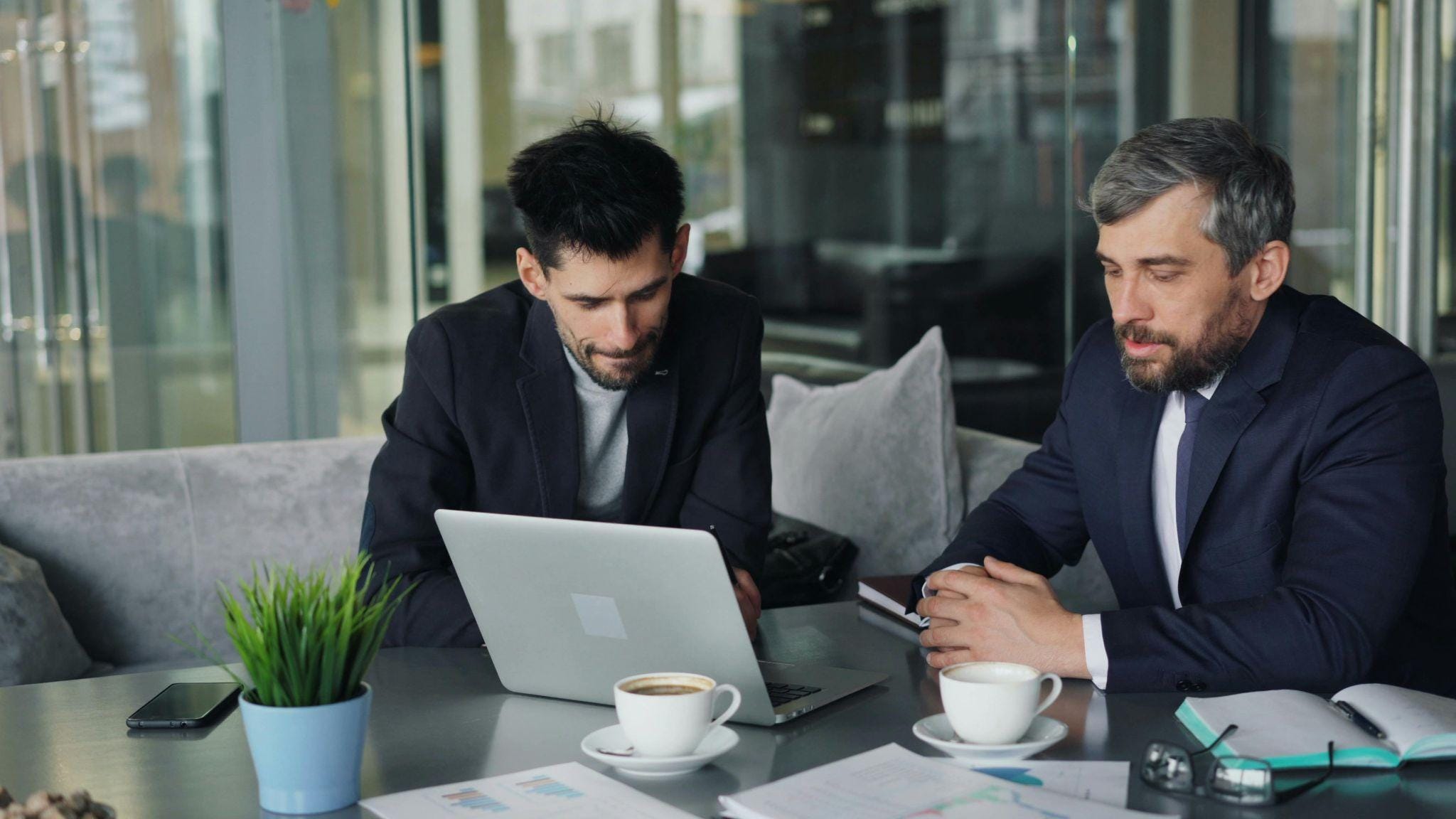 Deux hommes d'affaires en costume discutent devant un ordinateur portable dans un bureau moderne avec baies vitrées, café et plante verte sur la table.