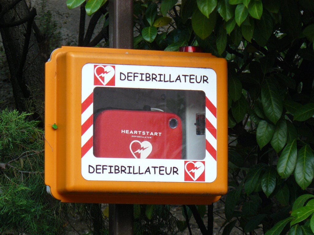 Arrêt cardiaque en entreprise : comment s’équiper et réagir efficacement ? 4 Boîtier défibrillateur automatisé externe orange fixé sur poteau, contenant un défibrillateur Heartstart rouge, entouré de lierre vert.