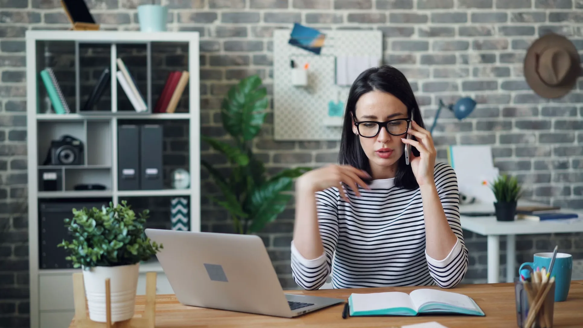 Quelles sont les principales pannes d’ordinateur et comment les identifier ? 3 Femme portant des lunettes en appel téléphonique devant son ordinateur portable dans un bureau moderne avec mur de briques, plantes et rangements.