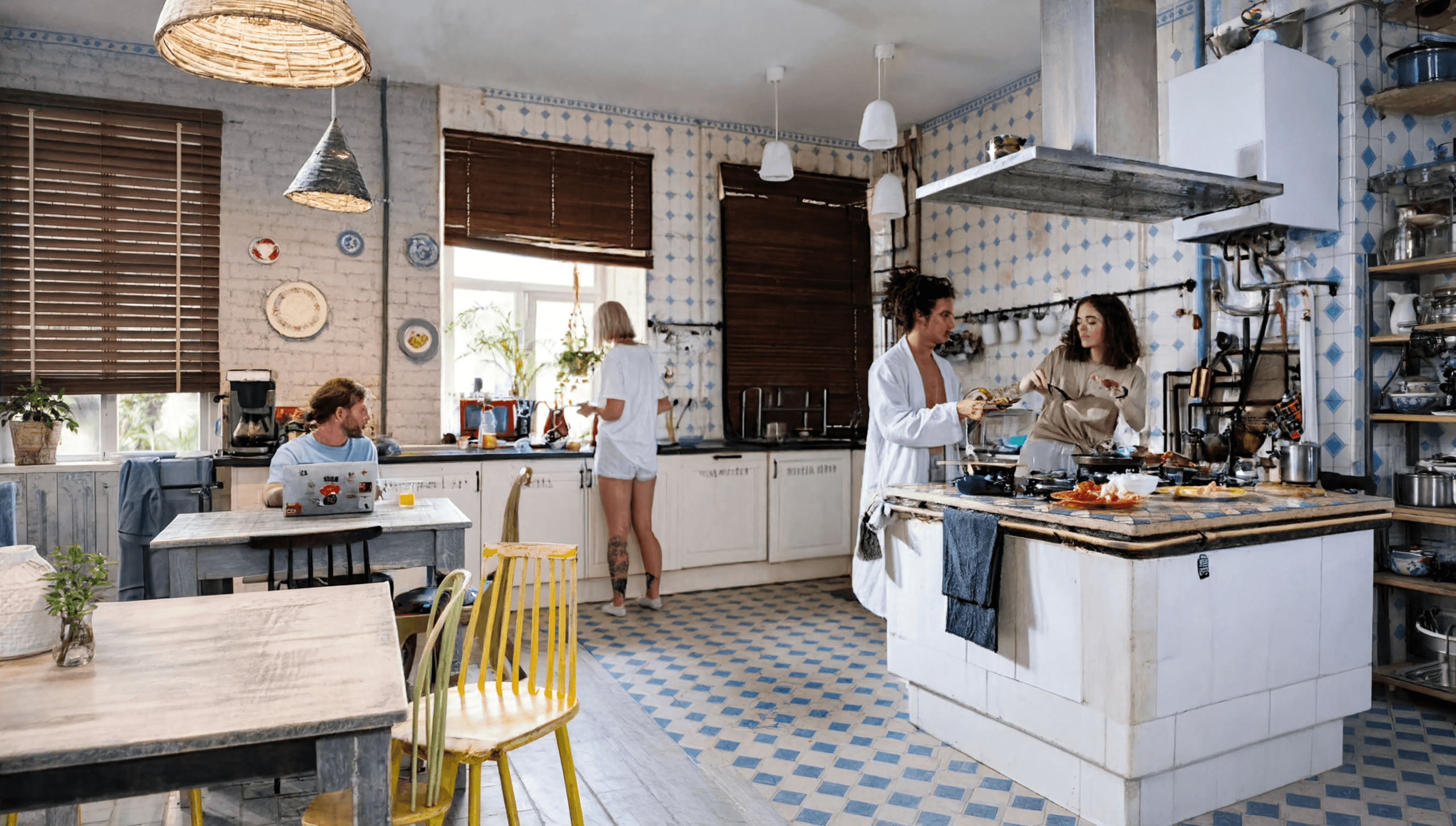 Cuisine spacieuse avec îlot central, carrelage bleu et blanc, plusieurs personnes cuisinant ensemble, armoires blanches, luminaires en paille, fenêtres avec volets bruns, étagères ouvertes.