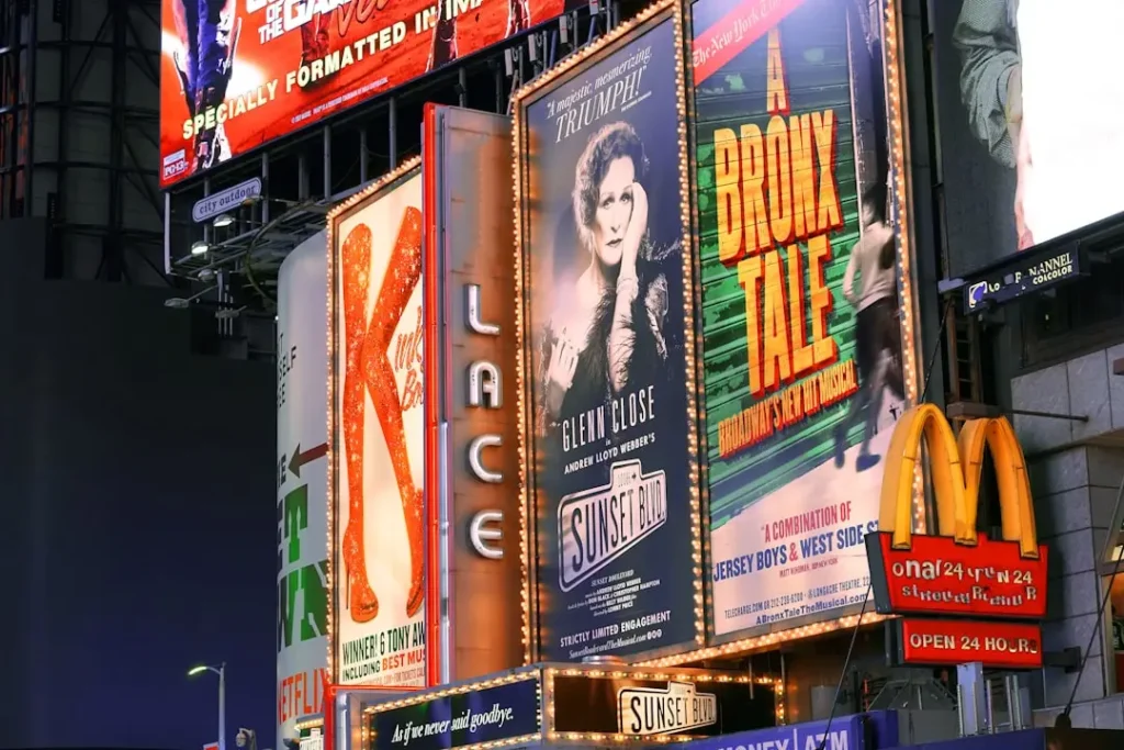 Façade urbaine nocturne avec affiches de spectacles musicaux et enseignes lumineuses, dont un roll-up publicitaire pour Sunset Boulevard et logos commerciaux illuminés.